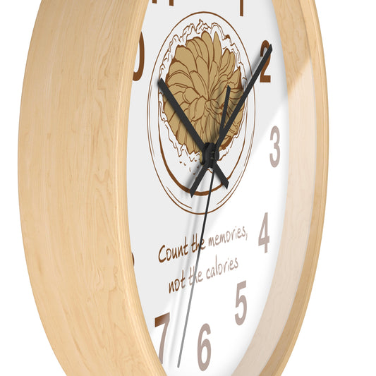 A close-up of a wall clock with a light natural wood frame, a white face, black hands, a tan floral illustration, and the phrase 'Count the memories, not the calories' on a white background.