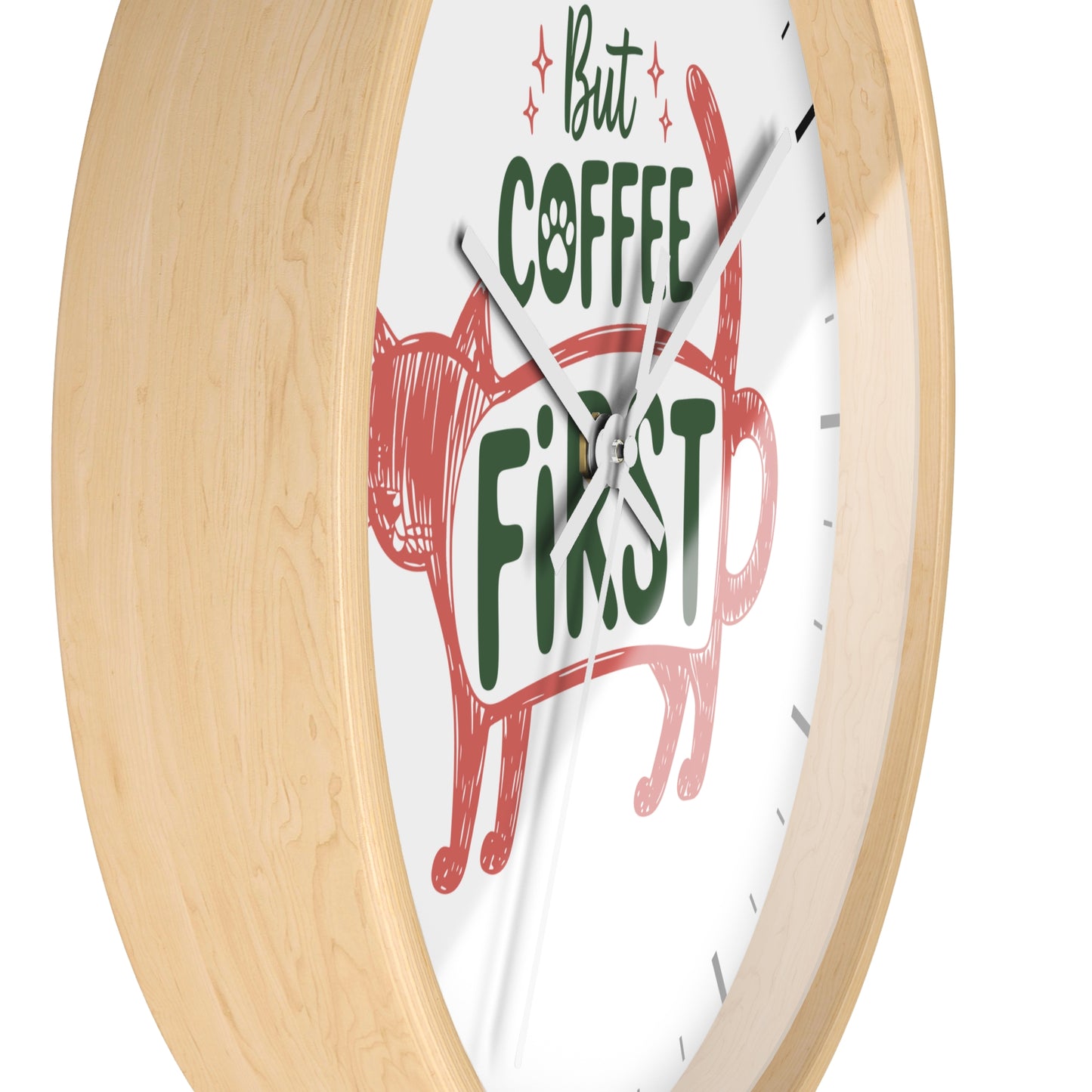 A close-up of a round wall clock with a light natural wood frame and a white face. The clock face features a red cat illustration and the phrase 'But COFFEE FIRST' in green and black lettering.