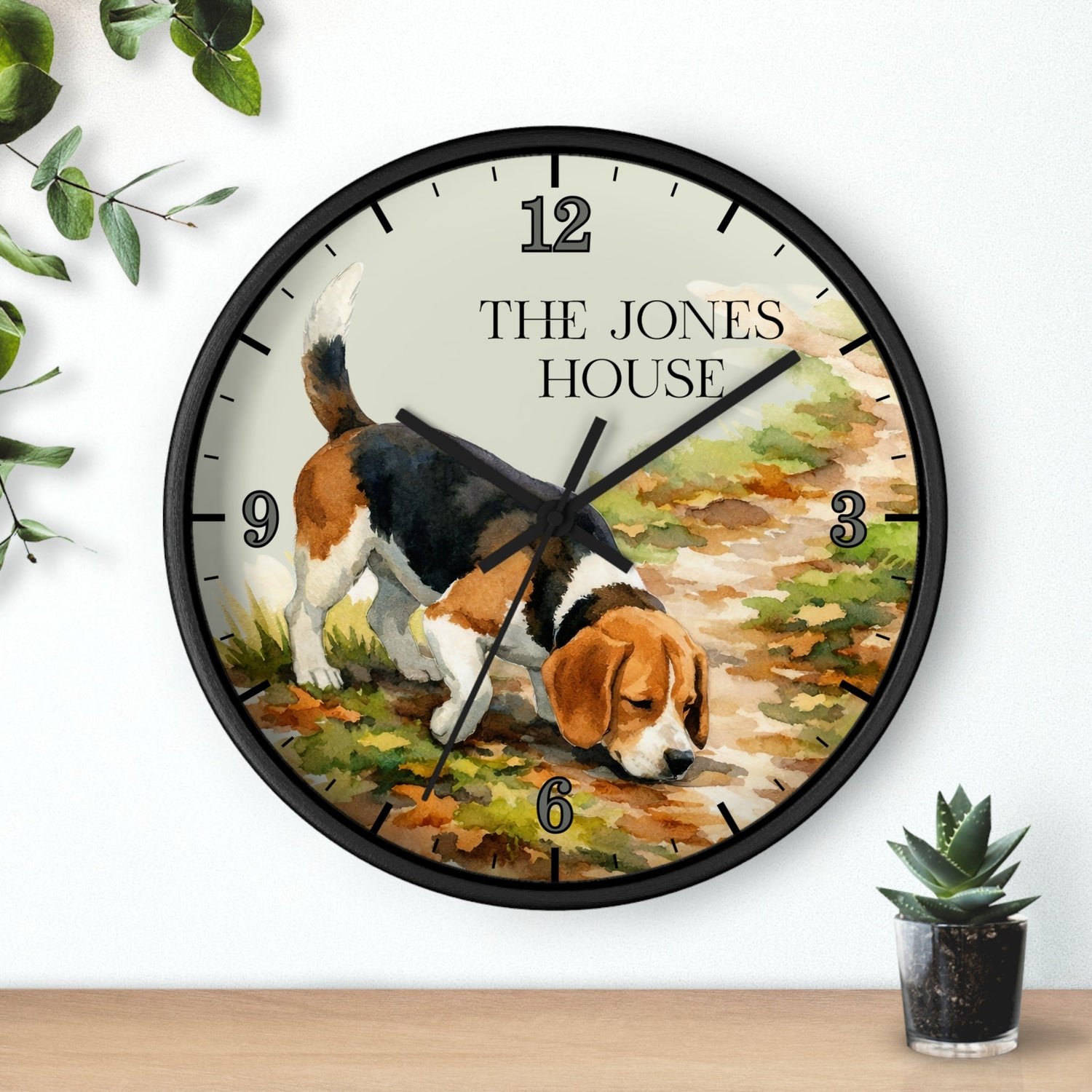 A round wall clock with a black frame and a printed illustration of a beagle dog sniffing the ground, with the text 'THE JONES HOUSE' at the top. The clock is mounted on a white wall above a wooden surface, with small green plants in the corners.