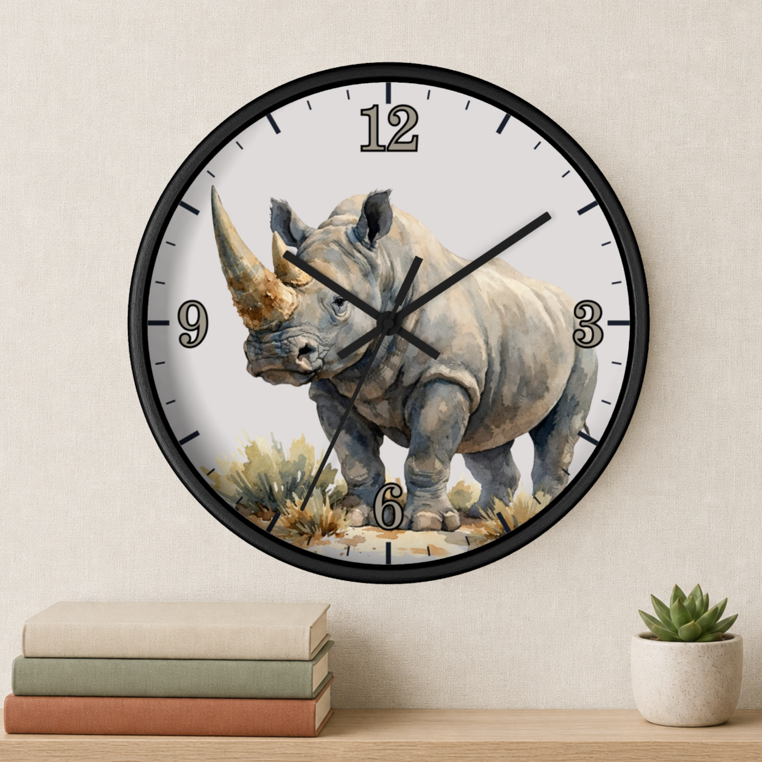 A round wall clock with a black frame and a watercolor-style rhinoceros illustration on the face, mounted on a light beige wall above a shelf with stacked books and a small potted succulent.