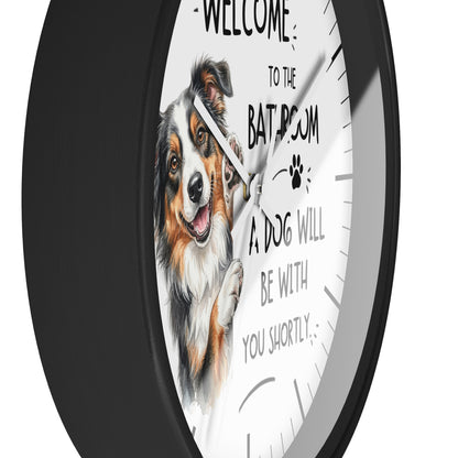 A round wall clock with a black frame and a white face featuring an illustration of a fluffy dog and the text 'WELCOME TO THE BATHROOM A DOG WILL BE WITH YOU SHORTLY'.