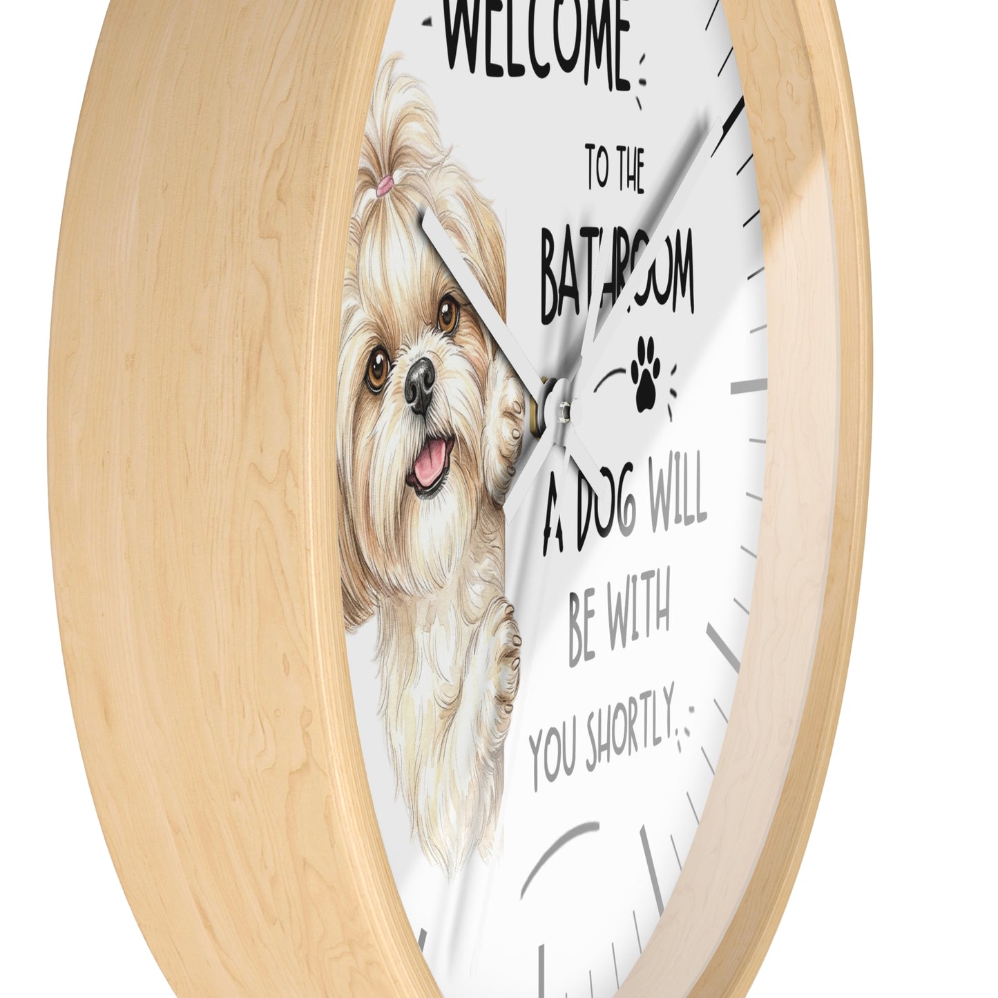 A round wall clock with a light natural wood frame and a white face. The clock features an illustration of a fluffy cream-colored dog on the left side and black text that reads 'WELCOME TO THE BATROOM A DOG WILL BE WITH YOU SHORTLY' along with a small paw print.