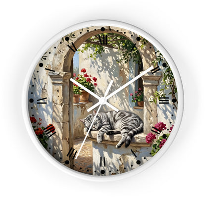 Round wall clock with a white frame and a printed scenic dial showing a gray tabby cat resting on a stone ledge beneath an arched doorway, surrounded by flowers and greenery. Black Roman numerals mark the hours, and white clock hands are visible against a plain white background.