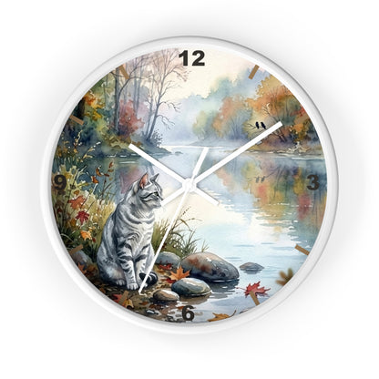 Round wall clock with a white frame and a printed scenic design featuring a gray tabby cat sitting by a river in an autumn landscape. The clock face includes black numerals and white hands on a white background.