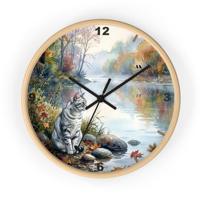 A round wall clock with a light natural wood frame, featuring a printed scenic illustration of a gray tabby cat sitting by a calm river in an autumn landscape. The clock face includes black Arabic numerals for hours and black clock hands.