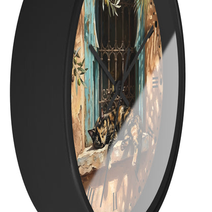 A round wall clock with a black frame and a printed scenic face showing a tortoiseshell cat resting on a windowsill in front of blue shutters and an iron gate.