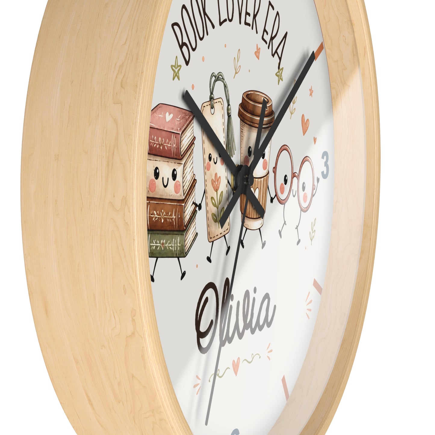 A round wall clock with a light natural wood frame and a white face featuring cute illustrated books, a coffee cup, and the text 'BOOK LOVER ERA' and 'Olivia'.