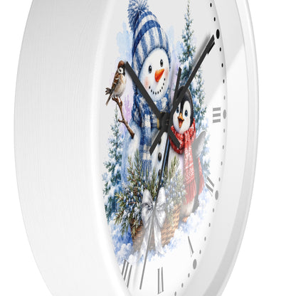 A white round wall clock with a winter-themed illustration on the face, featuring a snowman in a blue striped hat, a penguin in a red scarf, evergreen trees, and bird details. The clock is shown close-up against a white background.