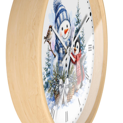 A round wall clock with a light natural wood frame and a white face featuring a winter illustration of a snowman, a penguin, birds, and evergreen trees. The clock is shown at an angle against a plain white background.