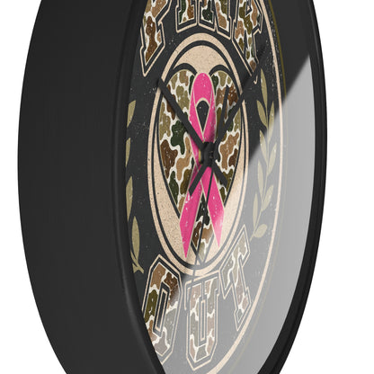 A close-up of a round wall clock with a black frame and a distressed graphic face. The design features a pink breast cancer ribbon over a camouflage heart, surrounded by olive green laurel branches in a tan block font.