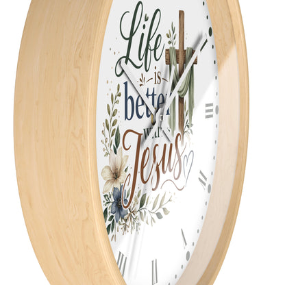 A round wall clock with a light natural wood frame and a white face featuring the phrase 'Life is better with Jesus' in decorative script and floral illustrations.
