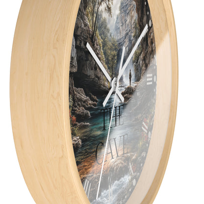Round wall clock with a light natural wood frame and a printed scenic face showing a cave interior with a river, trees, and the words “THE CAVE.” White clock hands are visible against a plain white background.