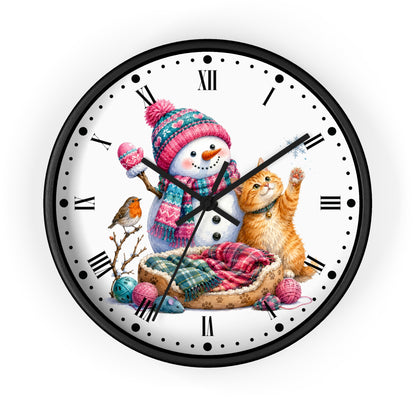 A round wall clock with a black frame and a white face featuring a festive winter illustration of a snowman in a pink knit hat, an orange tabby cat, a small bird, yarn balls, and a pet bed.