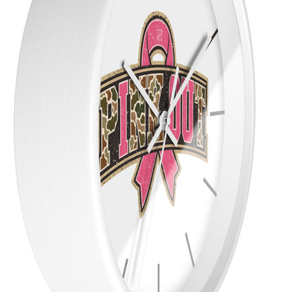 A close-up of a white wall clock face featuring a large pink ribbon and the word 'PIMP OUT' in a camouflage pattern, with silver hands against a clean white background.
