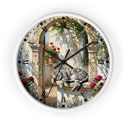 Round wall clock with a white frame and a printed scenic face showing a gray tabby cat sleeping on a stone ledge beneath an arched doorway with flowers and greenery. Roman numerals mark the hours, and the clock is photographed against a plain white background.