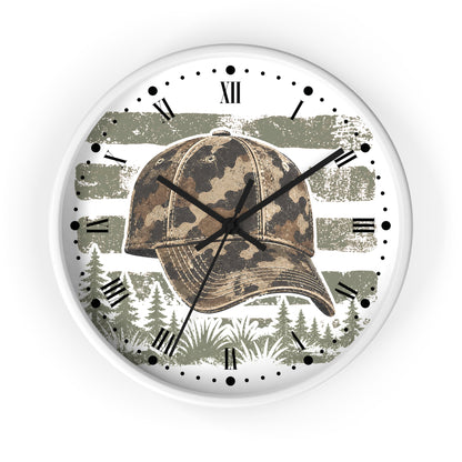 A round wall clock with a white frame and a printed design featuring a camouflage baseball cap over a distressed olive green and white American flag, with pine trees along the bottom. The clock face has black Roman numerals and black hands on a white background.