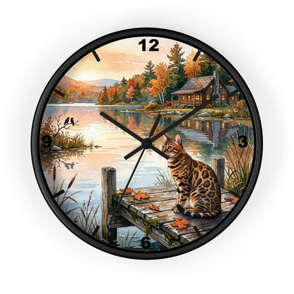 A round wall clock with a black frame and black numerals, featuring a scenic autumn lakeside illustration with a cat sitting on a dock. The clock is shown against a plain white background.