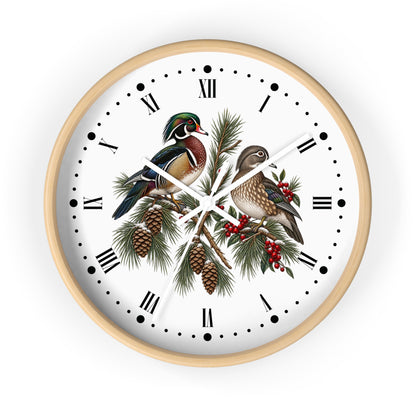A round wall clock with a light natural wood frame, featuring a white face decorated with an illustration of two wood ducks perched on pine branches with pinecones and red berries, set against a plain white background.