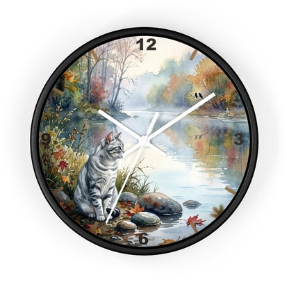 A round wall clock with a black frame and a printed scenic design featuring a gray tabby cat sitting by a river in autumn foliage. The clock face includes white clock hands, black numerals, and a light gray background around the product.