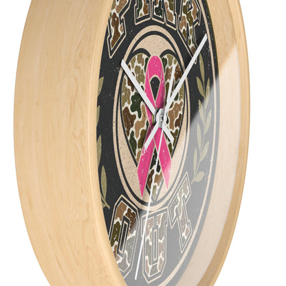 A close-up of a round wall clock with a light natural wood frame. The clock face features a black background with a central camouflage heart motif overlaid with a bright pink ribbon, and stylized text around the edge.
