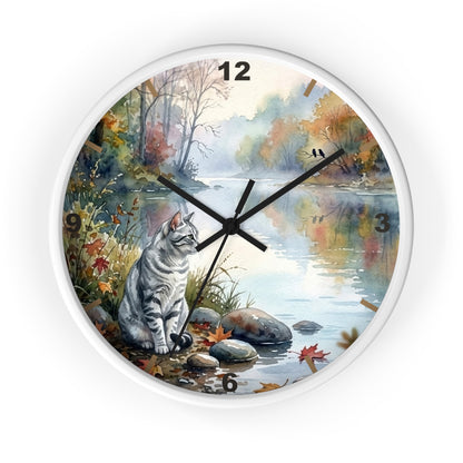 Round wall clock with a white frame and a printed scenic design featuring a gray tabby cat sitting by a river in autumn foliage. The clock face includes black Arabic numerals and black hands, shown against a plain white background.