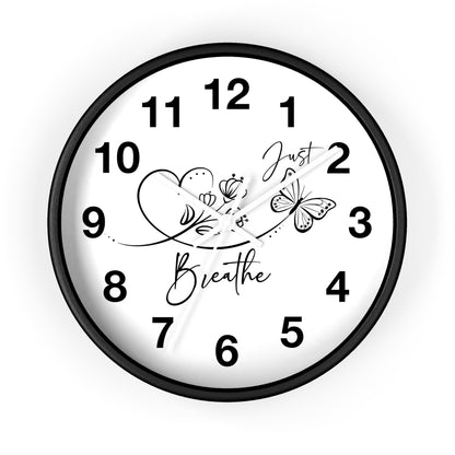 A round wall clock with a black frame and white face, featuring black Arabic numerals and decorative script and line art reading 'Just Breathe' with flowers and a butterfly. The clock is centered on a light gray background.