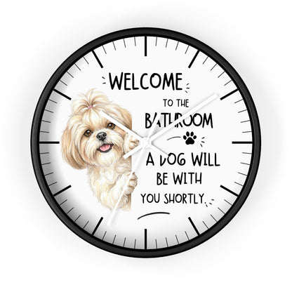 A round novelty wall clock with a black frame and white face, featuring a small fluffy dog illustration and the text 'WELCOME TO THE BATHROOM A DOG WILL BE WITH YOU SHORTLY.' on a light gray background.