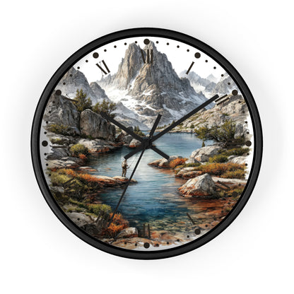 A round wall clock with a black frame and a scenic landscape print featuring a mountain range, rocky shoreline, blue water, and a small figure fishing. The clock face has black Roman numerals and black hands on a white background.