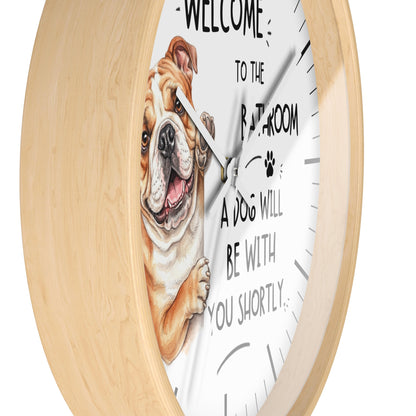 A round wall clock with a light natural wood frame and a white face featuring a cartoon bulldog and the text 'WELCOME TO THE BATHROOM A DOG WILL BE WITH YOU SHORTLY'.