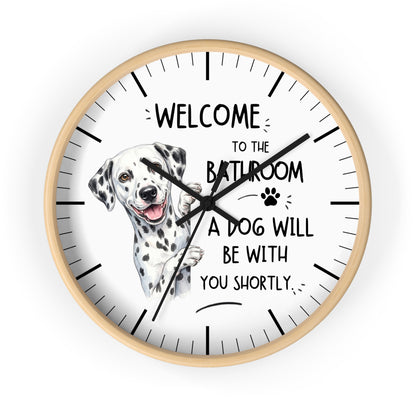 A round wall clock with a light wood frame and a white face, featuring a Dalmatian illustration and the text 'WELCOME TO THE BATHROOM A DOG WILL BE WITH YOU SHORTLY.' on a plain white background.