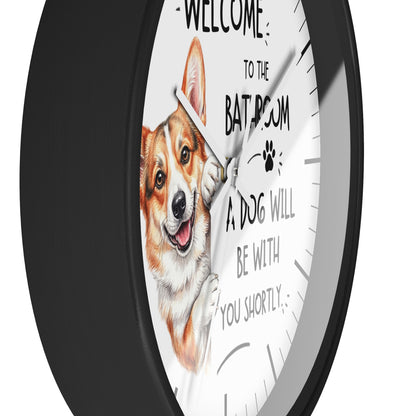 A round wall clock with a black frame and a white face, featuring a painted corgi illustration and the text 'WELCOME TO THE BATHROOM A DOG WILL BE WITH YOU SHORTLY'.