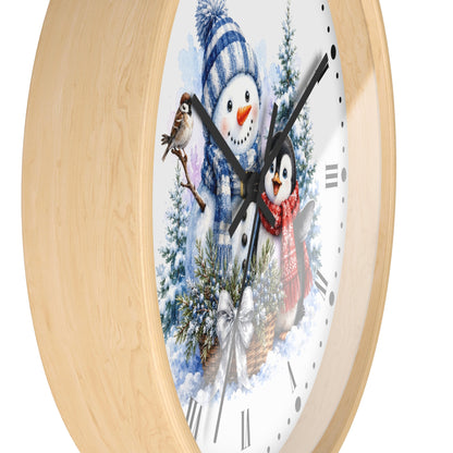 Round wall clock with a light natural wood frame and a white face featuring a winter holiday illustration of a snowman, a penguin, evergreen trees, and decorative clock hands.
