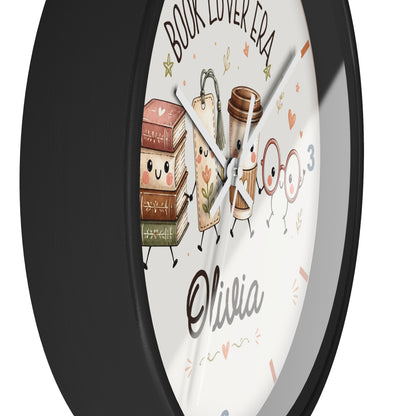 A round wall clock with a black frame and a white face featuring cute illustrated books, a coffee cup, glasses, and the text 'BOOK LOVER ERA' and 'Olivia'.