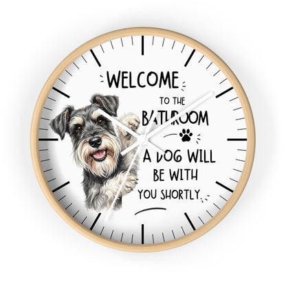 A round wall clock with a light wood frame and a white face, featuring a sketch of a gray and white schnauzer dog and the humorous text 'WELCOME TO THE BATHROOM A DOG WILL BE WITH YOU SHORTLY.' The clock is shown against a plain white background.