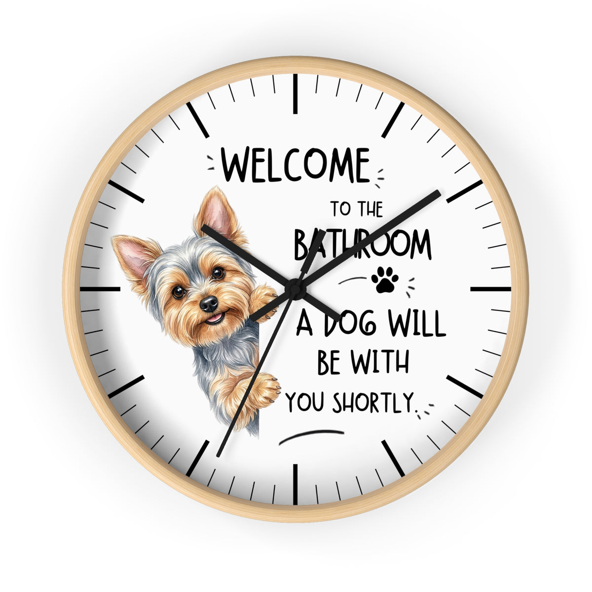A round wall clock with a light wood frame and a white face, featuring a cartoon illustration of a Yorkshire Terrier and the humorous text 'WELCOME TO THE BATHROOM A DOG WILL BE WITH YOU SHORTLY.' The clock hands are black and the hour markers are black lines.