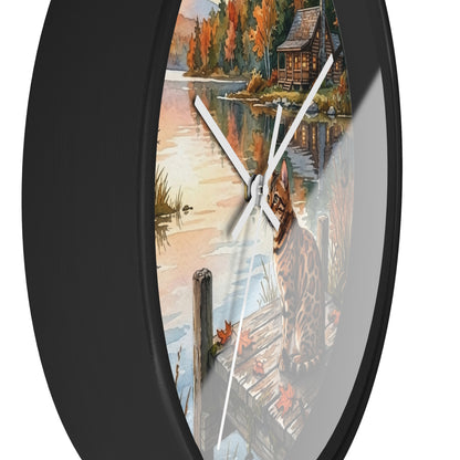A round wall clock with a black frame and white hands, featuring a scenic autumn lakeside illustration with a cabin, dock, and a spotted cat on the clock face.