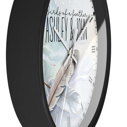 A close-up of a round wall clock with a matte black frame and a white dial featuring a feather illustration and the text 'birds of a leather ASHLEY & JOHN'.