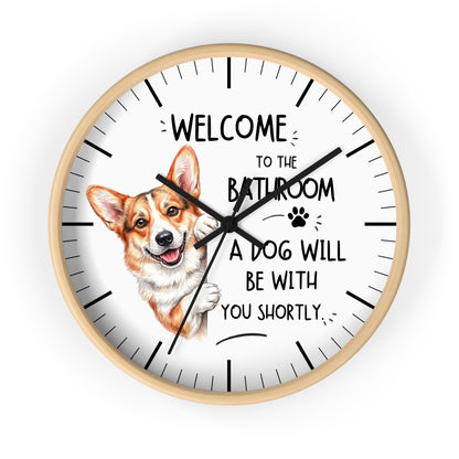 A round wall clock with a light wood frame and a white face, featuring a cartoon Corgi illustration and the text 'WELCOME TO THE BATHROOM A DOG WILL BE WITH YOU SHORTLY.' on a plain white background.