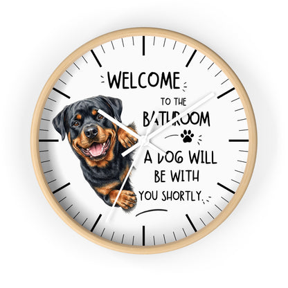 A round wall clock with a light wood frame and a white face, featuring a Rottweiler dog illustration and the text 'WELCOME TO THE BATHROOM A DOG WILL BE WITH YOU SHORTLY.' on a plain white background.