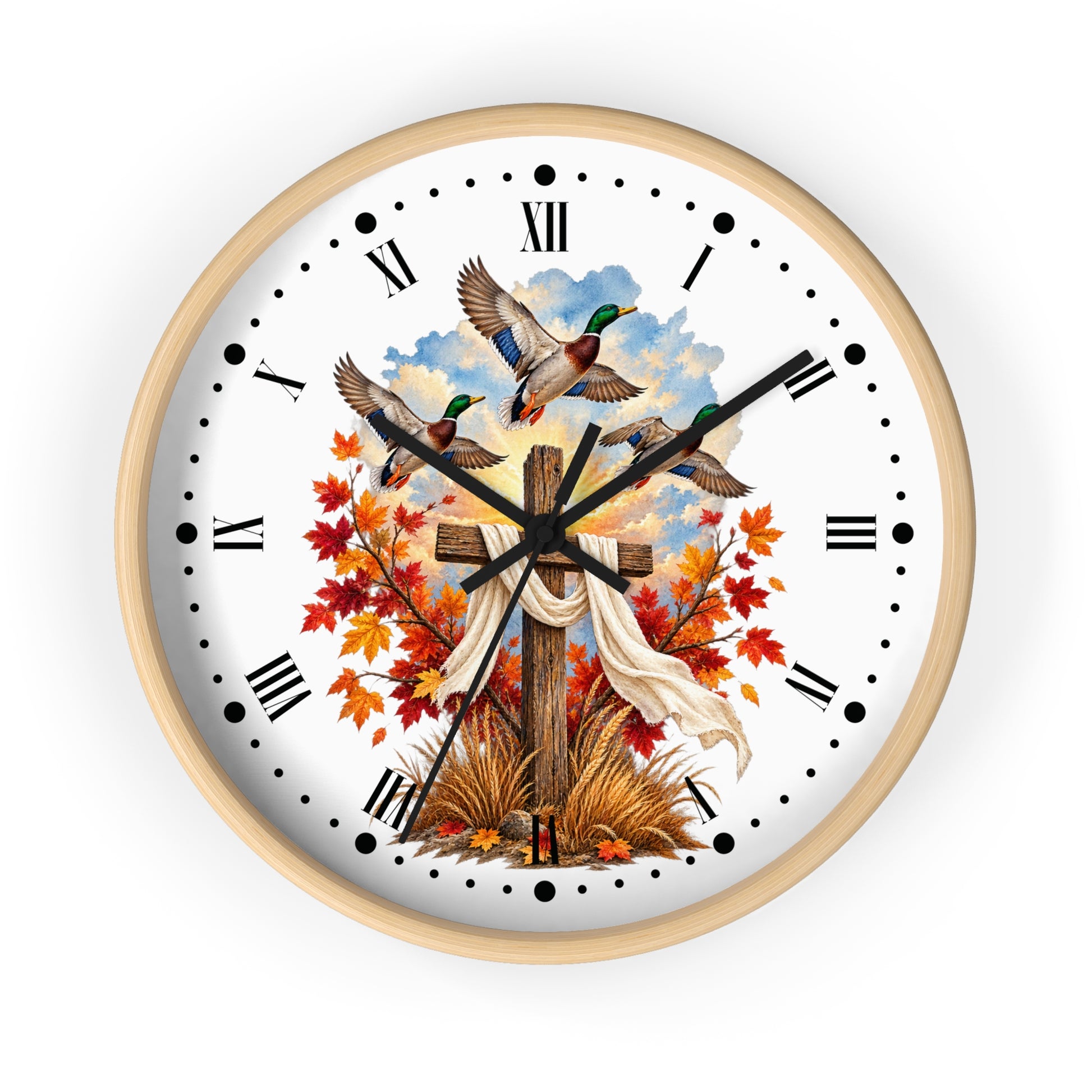 A round wall clock with a light natural wood frame and a white face, featuring a printed design of three mallards flying above a wooden cross draped with a white cloth, surrounded by red and orange autumn leaves and grass. The clock has black Roman numerals and hands on a clean white background.