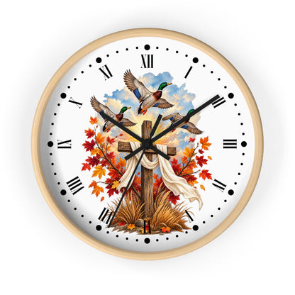 A round wall clock with a light natural wood frame and a white face, featuring a printed design of three mallards flying above a wooden cross draped with a white cloth, surrounded by red and orange autumn leaves and grass. The clock has black Roman numerals and hands on a clean white background.
