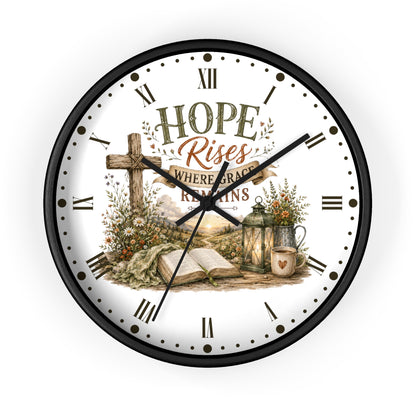 A round wall clock with a black frame and a white face featuring a Christian-themed illustration and the words 'HOPE Rises WHERE GRACE REMAINS.' The clock includes Roman numerals, black hands, and decorative floral and rustic scene details on the dial.
