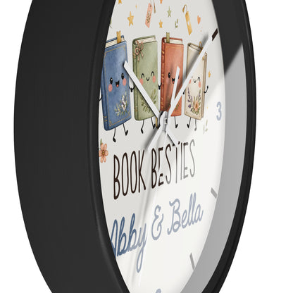 A round wall clock with a black frame and a white face featuring four cute illustrated books with faces and the text 'BOOK BESTIES Abby & Bella'.
