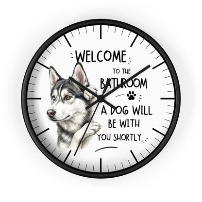 A round wall clock with a black frame and a white face, featuring a sketch of a husky dog and the text 'WELCOME TO THE BATHROOM A DOG WILL BE WITH YOU SHORTLY.' on a light gray background.