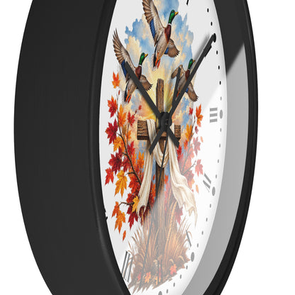A close-up of a wall clock with a black frame and a white face featuring a hunting cross, mallards, autumn leaves, and a white banner.