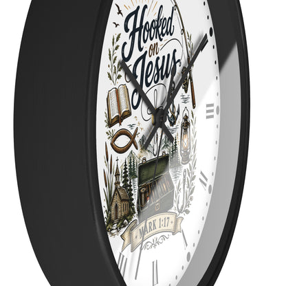 A close-up of a wall clock with a black frame and a white face featuring the text 'Hooked on Jesus' and religious illustrations including an open book, fish symbol, church, and lantern.