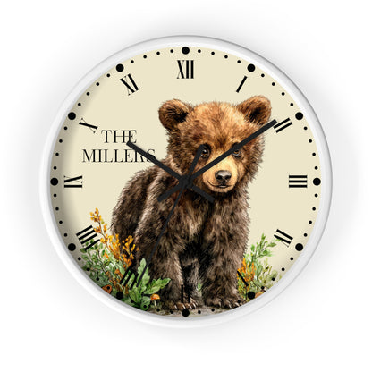 A round wall clock with a white frame and a cream-colored face featuring an illustration of a brown bear cub standing among green plants and yellow flowers. The clock has black Roman numerals and black hands, with the text 'THE MILLERS' printed on the left side.