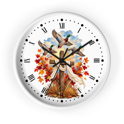 A round wall clock with a white frame and a printed autumn hunting scene on the face. The design shows three mallards flying above a wooden cross draped with a white cloth, surrounded by red and orange autumn leaves, grass, and a blue sky.