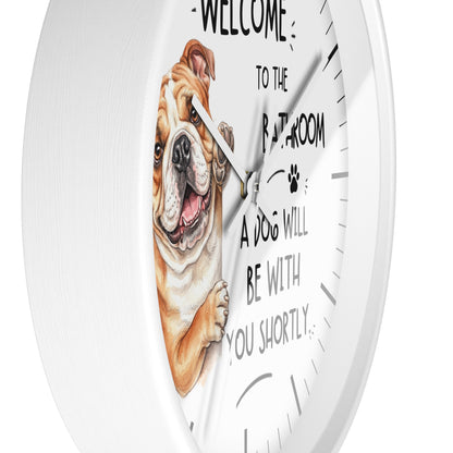 A white round wall clock with a humorous pet message printed on the face: 'WELCOME TO THE BATHROOM A DOG WILL BE WITH YOU SHORTLY.' The clock face features a detailed illustration of a brown and white bulldog peeking in from the left.