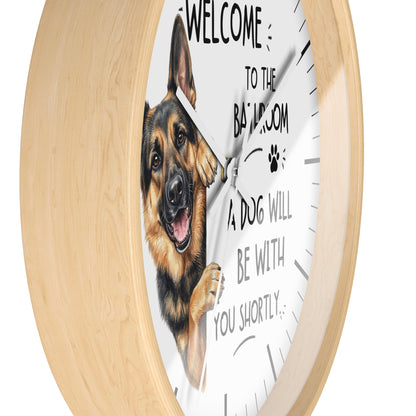 A round wall clock with a light natural wood frame and a white face featuring a German Shepherd illustration and the text 'WELCOME TO THE BATHROOM A DOG WILL BE WITH YOU SHORTLY'